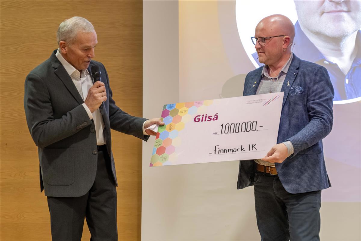 Thomas Møllman fra FeFo overrekker en stor symbolsk sjekk til Geir Knutsen fra Finnmark idrettskrets under Idrettshelga i Alta. De smiler og holder sjekken sammen foran publikum. - Klikk for stort bilde